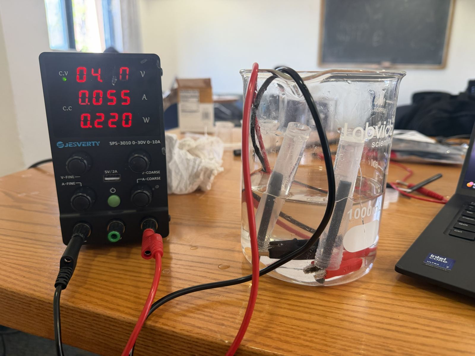 Bench power supply reading 4.0 volts and 0.055 amps, wired with red and black leads to two graphite electrodes in a beaker of electrolyte with inverted collection tubes.